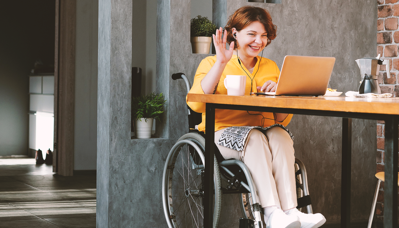 Women in a wheel chair wearing a bright yellow shirt and waves to the laptop screen.