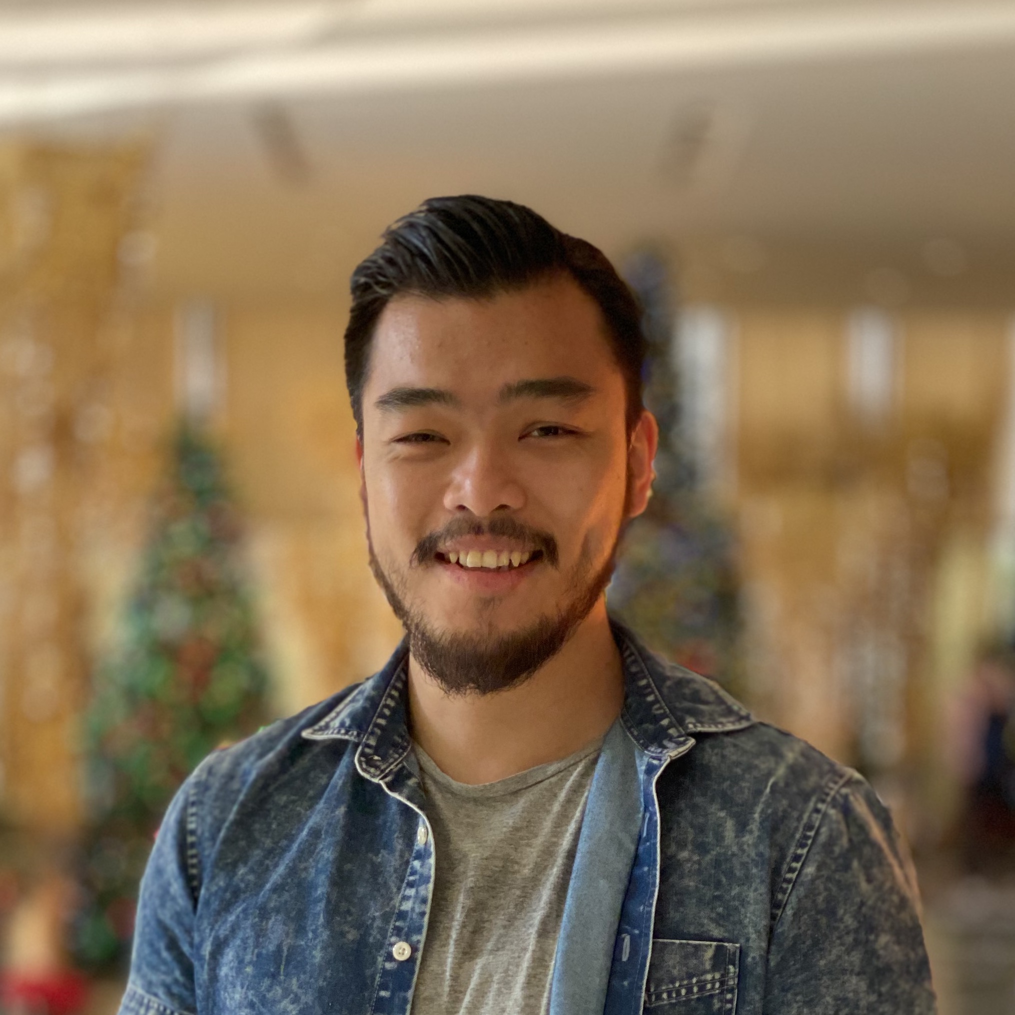 Aaron Chu, an east asian man with a beard and mustache smiling into the camera in front of a blurred background with golden architectural elements and christmas trees. He is wearing a grey t-shirt and a denim shirt.