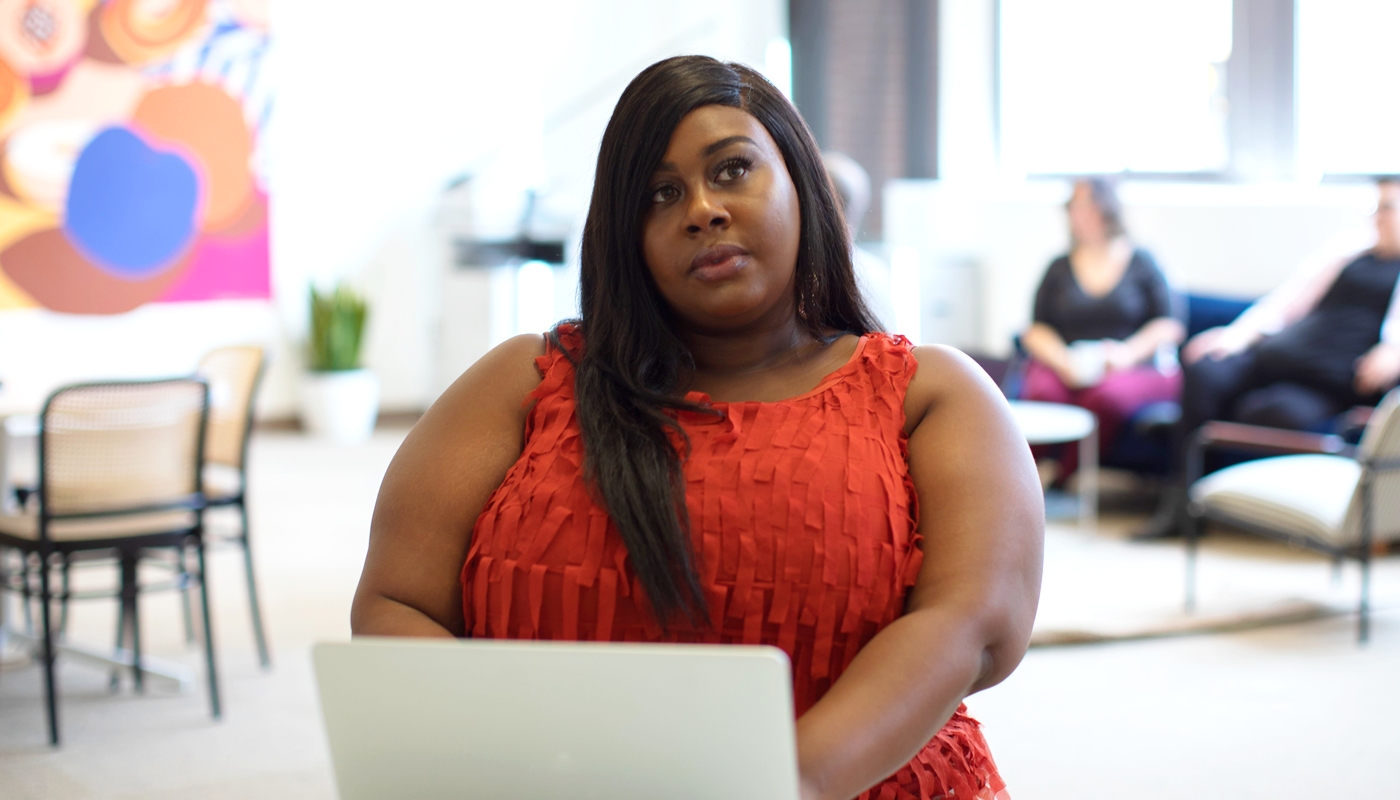 A Black woman sits in an office space working at her laptop, looking off into the distance.