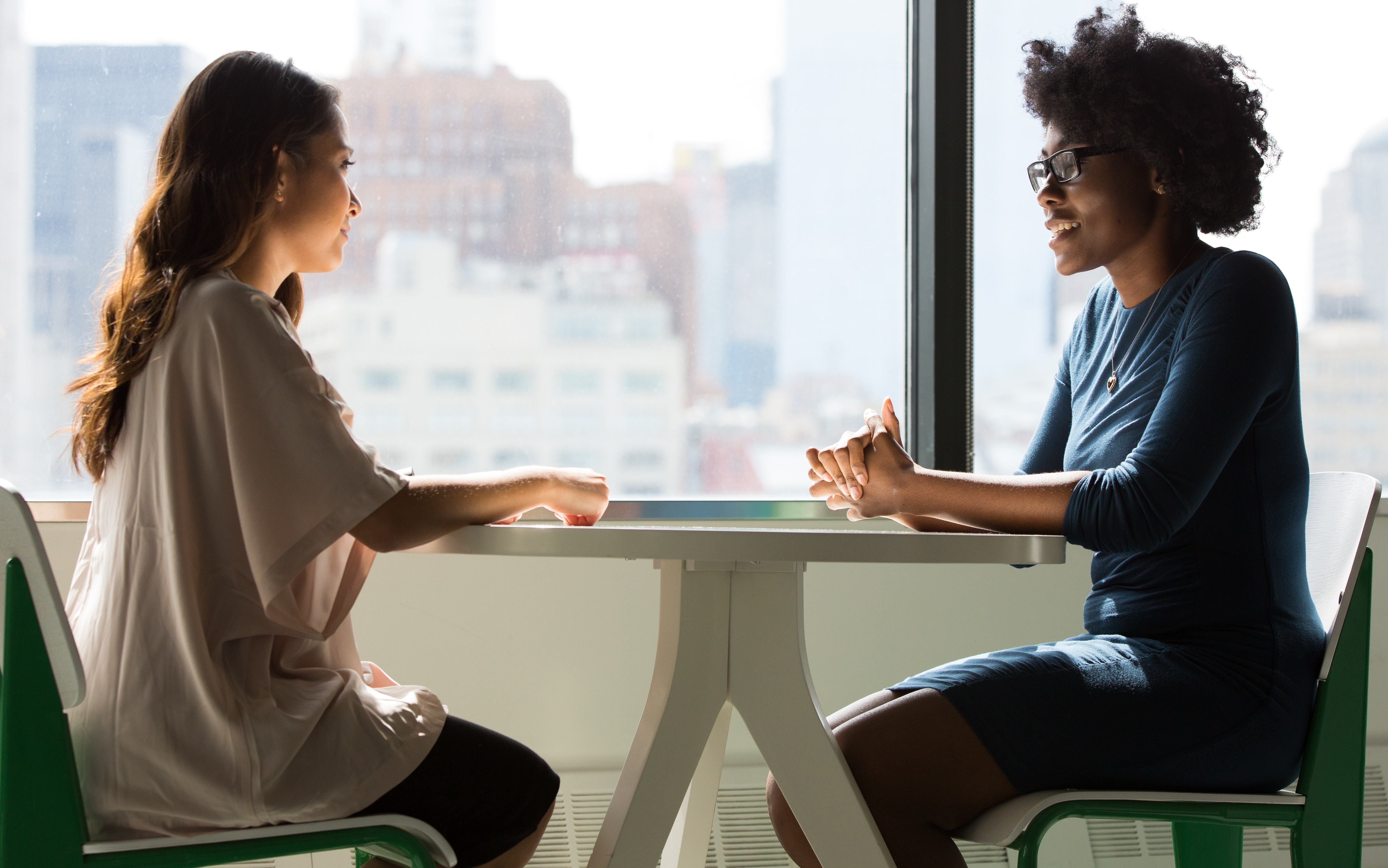 Two women of color sit an office table talking. We see their arms rest of the table before them and a city skyline through the window.
